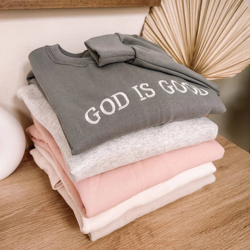 Stack of folded sweatshirts with 'GOD IS GOOD' text on a wooden surface.