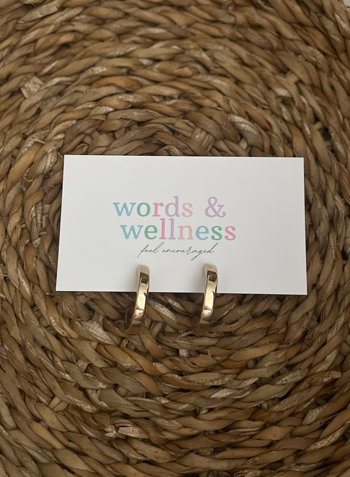 Gold hoop earrings on a 'words & wellness' card against a woven mat background