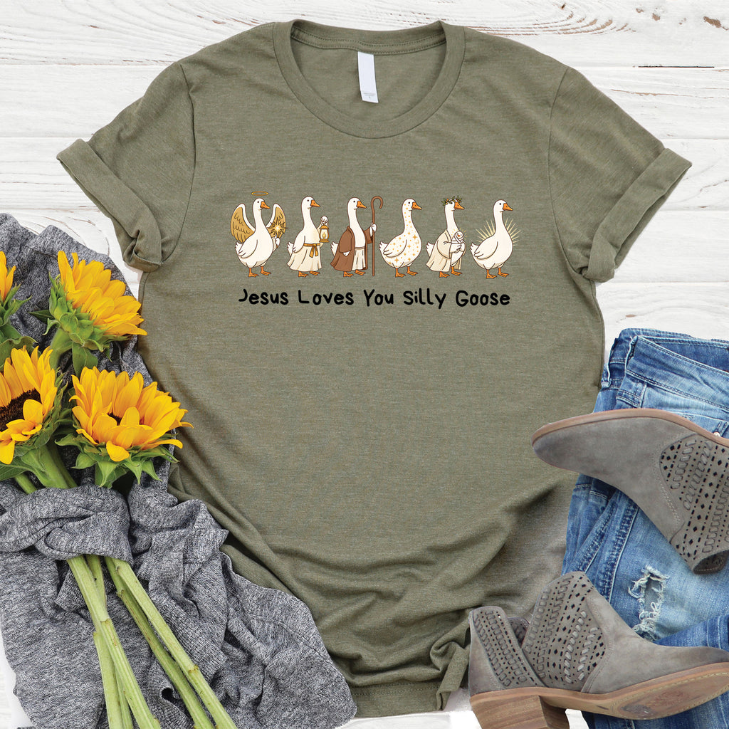 Green t-shirt with 'Jesus Loves You Silly Goose' graphic on a wooden surface with sunflowers and jeans.