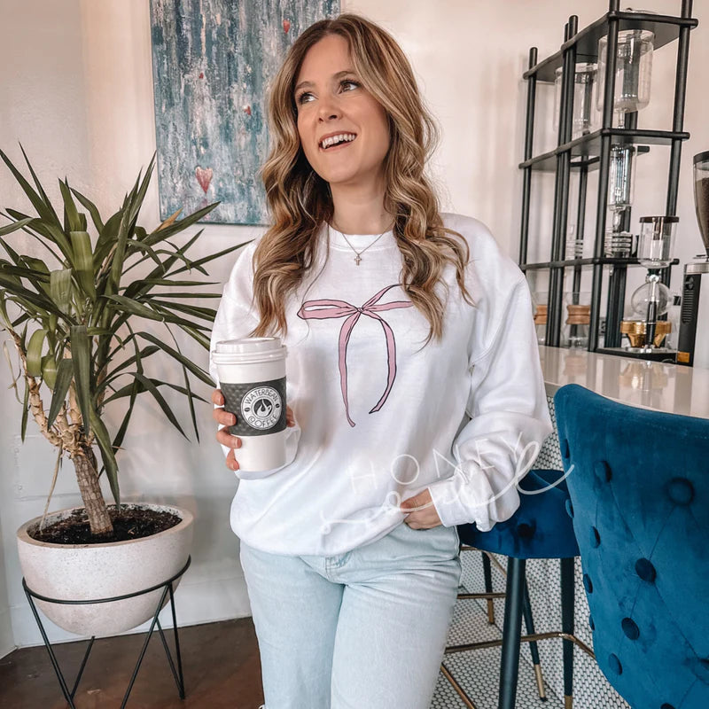Woman in a white sweatshirt with a pink design, holding a coffee cup, standing in a modern living room.